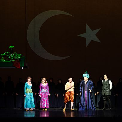 Szene aus Entf&uuml;hrung aus dem Serail: Menschen stehen vor einer &uuml;bergro&szlig;en t&uuml;rkischen Flagge