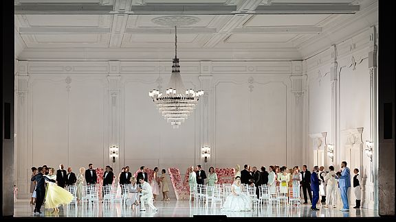 Foto einer Szene aus Semele: Eine Hochzeitsgesellschaft in einem gro&szlig;en Saal mit einem Kronleuchter an der Decke