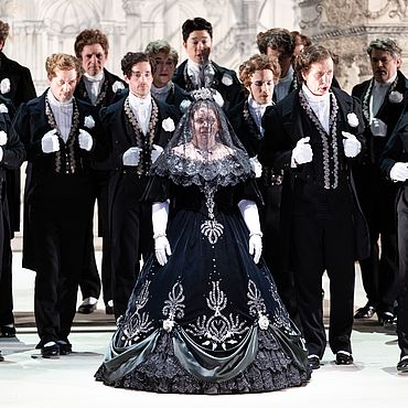 Szenenbild: La Cenerentola; Eine Frau in einem schwarzen Kleid umgeben von M&auml;nnern
