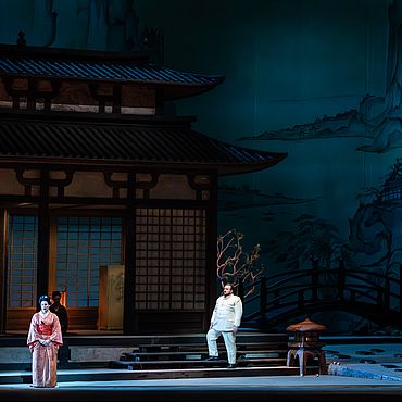 Szenenbild Madama Butterfly: Eine Frau in traditionellem japanischen Gewand steht vor einem Haus, ein Mann in uniform hinter ihr