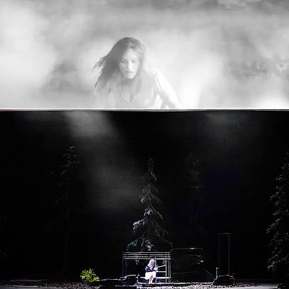 Sieglinde is in the forest, in the burned-down cabin where she grew up; on the screen above the stage, her mother can be seen in the flames.