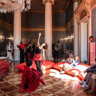 People sit on large red cushions and watch the dancers